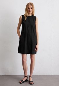 Schwarzes ärmelloses Kleid mit geradem Schnitt, rundem Ausschnitt, Seitentaschen und glatter Textur. Getragen mit schwarzen flachen Sandalen.
