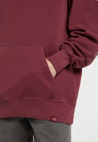 Osoba s rukou v kapse vínového svetru, který má žebrované manžety a malou červenou nášivku Dickies.