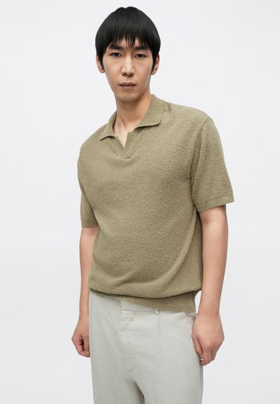 Κοντομάνικο πόλο μπλουζάκι σε ελαιί πράσινο με ανάγλυφη υφή, με μερικό γιακά και άνετη γραμμή, συνδυασμένο με ανοιχτό μπεζ παντελόνι.