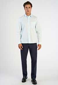 Lichtblauw overhemd met lange mouwen, knoopsluiting aan de voorkant en borstzak, gecombineerd met donkerblauwe broek en zwarte schoenen.