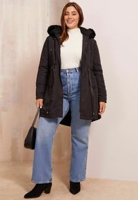 Fekete pehelydzseki, prémes kapucnival, húzózsinóros derékkal, cipzáras és gombos záródással, világos pulóver és magasderekú farmer fölött viselve.