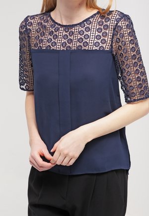 Marineblauwe top met doorschijnende korte mouwen en kant boven op de borst met cirkelvormige bloemmotieven, gecombineerd met een zwarte broek.