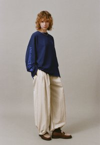 Felpa blu navy con la scritta "TO STAND OUT" stampata in blu sulla manica, abbinata a pantaloni beige a gamba larga e texture e mocassini marroni.