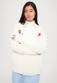 Vit stickad tröja med färgglad broderi av karaktärer i randiga skjortor. Modellen bär en ljusblå, randig skjorta med krage under.