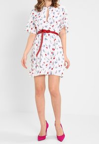 Kvinna klädd i en vit blommig klänning med röd bälte och rosa högklackade skor, stående mot en ljus enfärgad bakgrund.