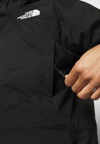 Zwarte jas van een gestructureerde stof, met een ritszak en het The North Face-logo geborduurd op de borst.