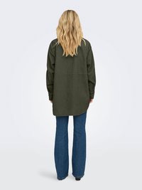 Camisa de veludo cotelê verde com mangas longas, barra arredondada e detalhe na parte de trás, combinada com jeans azul de corte flare.