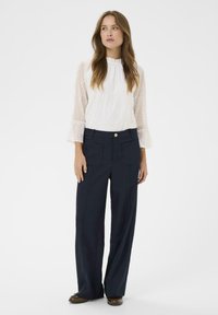 Blouse blanche avec un motif en œillets et un col froncé, associée à un pantalon large bleu marine avec des poches avant et un bouton doré.