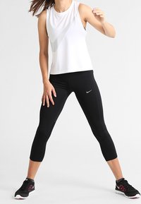 Mujer vistiendo una blusa sin mangas blanca, leggings cortos negros y zapatillas negras en una postura amplia frente a un fondo blanco liso.