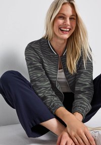 Veste bomber à motif zèbre en vert foncé et noir, avec une fermeture éclair à l'avant et des poignets côtelés. Associée à un pantalon ample bleu marine.