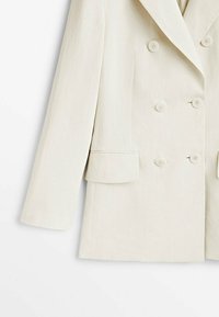 Gros plan d'un blazer croisé couleur crème avec six boutons et une poche à rabat sur le côté gauche.