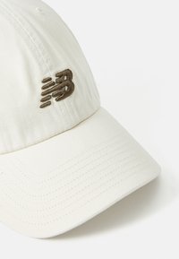 Casquette de baseball blanche avec visière incurvée et logo brodé marron sur le panneau avant, sur fond uni.
