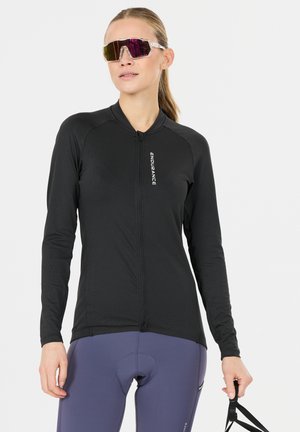 Femme portant un maillot de cyclisme noir à manches longues et un short de cyclisme rembourré violet, tenant une sangle noire, portant des lunettes de sport réfléchissantes.