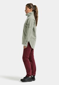 Bluza polarowa w jasnym zielonym kolorze, z wysokim kołnierzem i bocznym rozcięciem, zestawiona z bordowymi spodniami i czarnymi butami turystycznymi. Posiada kieszeń na lewym ramieniu.