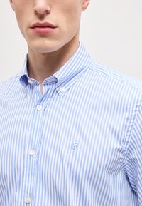 Κοντινό πλάνο ατόμου που φοράει πουκάμισο με κάθετες ρίγες σε ανοιχτό μπλε και λευκό χρώμα, με μικρό κεντημένο λογότυπο στο στήθος.