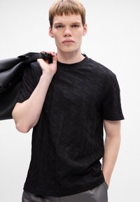 Czarny T-shirt z teksturowanym geometrycznym wzorem, okrągłym dekoltem, krótkimi rękawami i luźnym krojem, w połączeniu z czarną torbą noszoną na ramieniu.