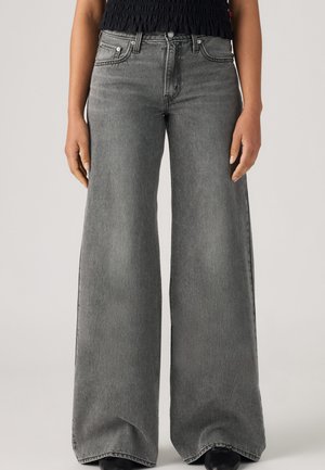 Personne portant un jean gris taille haute à jambes larges et un top noir, debout, les mains détendues le long du corps, sur un fond uni.