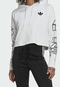 White cropped hoodie with long sleeves featuring a black Adidas logo on the chest and bold black text along the sleeves. Cotton blend fabric.