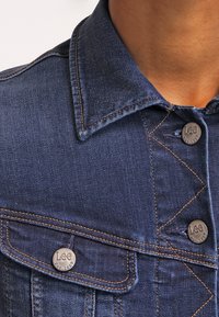 Mörk denimjacka med spetsig krage, silverfärgade knappar med "Lee"-logotyp och kontrasterande orangea sömmar.