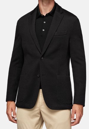Man die een zwarte tweeknopssblazer draagt over een zwart poloshirt en beige broek, staand met ontspannen armen langs het lichaam.