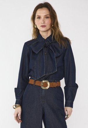 Donna che indossa una camicetta di denim scuro con fiocco oversize, pantaloni di denim abbinati, cintura marrone con grande fibbia dorata e gioielli dorati su sfondo bianco.