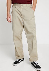 Beige Chinos mit Seitentaschen, Gürtelschlaufen und geradem Bein Design. Kombiniert mit schwarzen Sneakers mit weißen Akzenten.