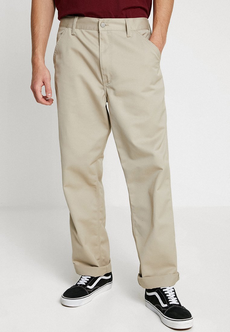 Beige Chinos mit Seitentaschen, Gürtelschlaufen und geradem Bein Design. Kombiniert mit schwarzen Sneakers mit weißen Akzenten.