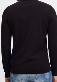 Homme portant un pull noir à manches longues et un jean bleu, vu de dos sur un fond uni.