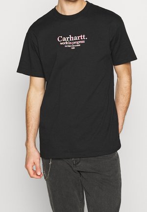 T-shirt nera in cotone con maniche corte, caratterizzata da un testo grafico bianco e arancione. Abbinata a pantaloni grigi e un accessorio a catena.