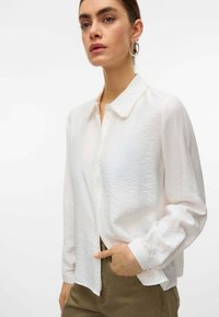 Camicetta bianca con finitura testurizzata, dettagli in perla sul colletto e maniche lunghe. Indossata con pantaloni color marrone chiaro.