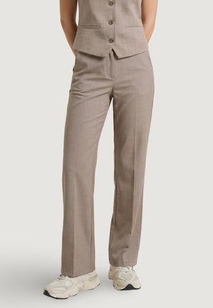 Persona con pantalones beige de pierna recta y chaleco abotonado a juego con zapatillas blancas sobre un fondo liso.