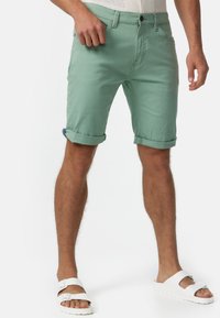Mintgrüne Shorts aus Baumwolle mit umgeschlagenen Säumen, ausgestattet mit frontalen Taschen und einem Knopfverschluss. Getragen mit weißen Sandalen.