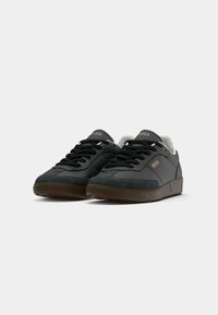 Paire de baskets basses noires avec semelles marron, lacets noirs et marquage doré « BOSS » sur le côté et la languette, sur un fond blanc uni.