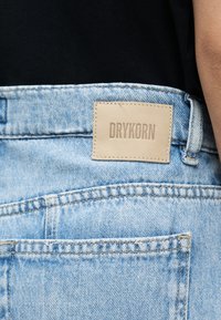 Lys blå jeans i denim med baklomme og sydd beige lærmerke med teksten "DRYKORN", brukt sammen med en svart topp.
