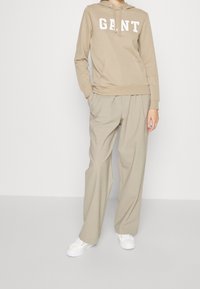 Bežová mikina s kapucí a bílým logem "GANT", v kombinaci s volnými bežovými kalhotami a bílými teniskami. Hladká textura, ležérní design.