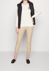 Svart vadderad väst över en vit kabelstickad tröja, beige beskurna byxor och svarta loafers. Har logotyp på tröjan.