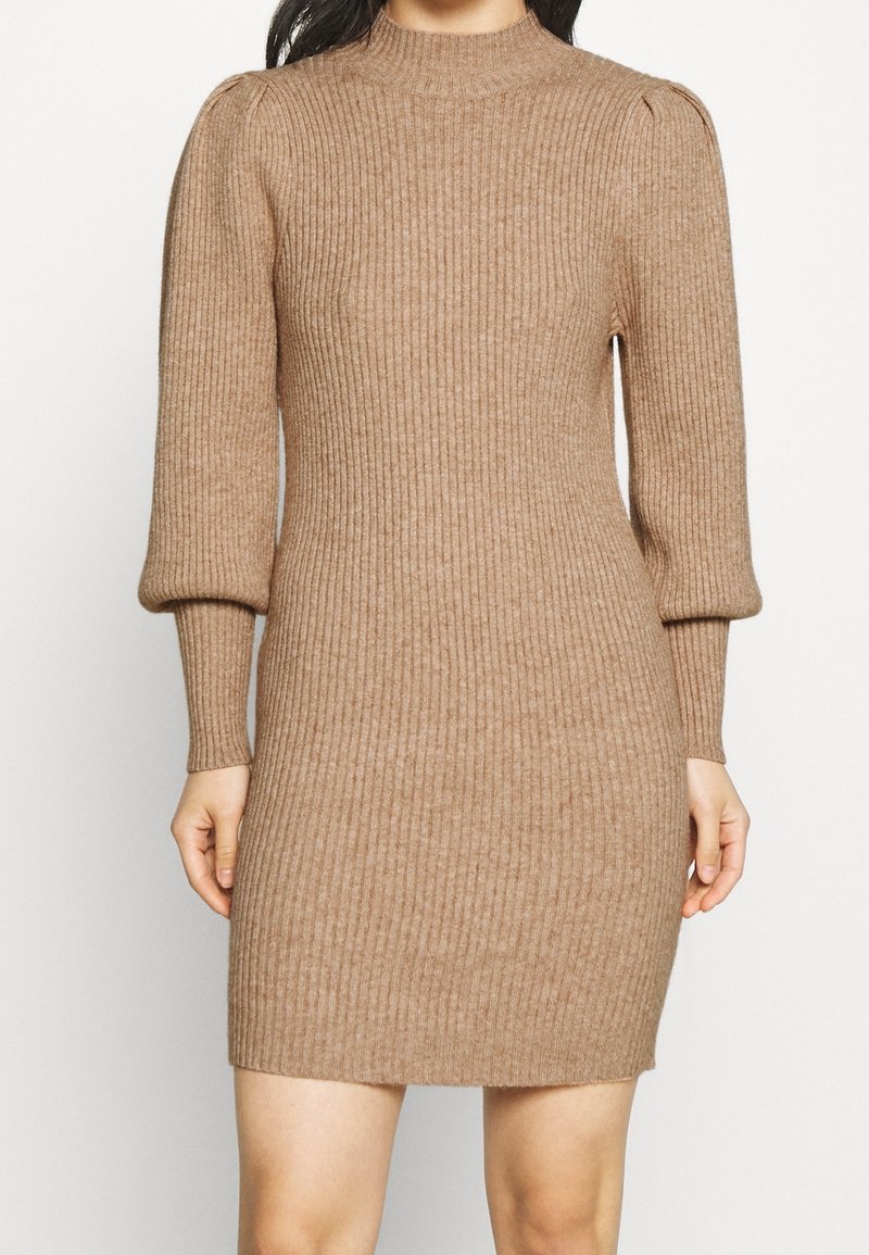 Ribbad stickad klänning i beige, med hög hals, puffärmar och en åtsittande silhuett som slutar ovanför knäna. Slät textur genom hela plagget.