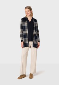 Marinblå och krämfärgad rutig cardigan med öppen framsida, bärs över en svart topp. Kombineras med beige vida byxor och bruna skor.