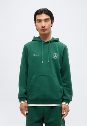 Jeune homme portant un sweat à capuche vert foncé avec le texte blanc "It's on us." et un logo circulaire sur la poitrine, debout devant un fond clair uni.