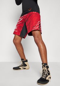 Shorts à motif rouge et noir en tissu lisse, avec des accents blancs et des fentes sur les côtés, associés à des chaussures montantes noires et beiges.