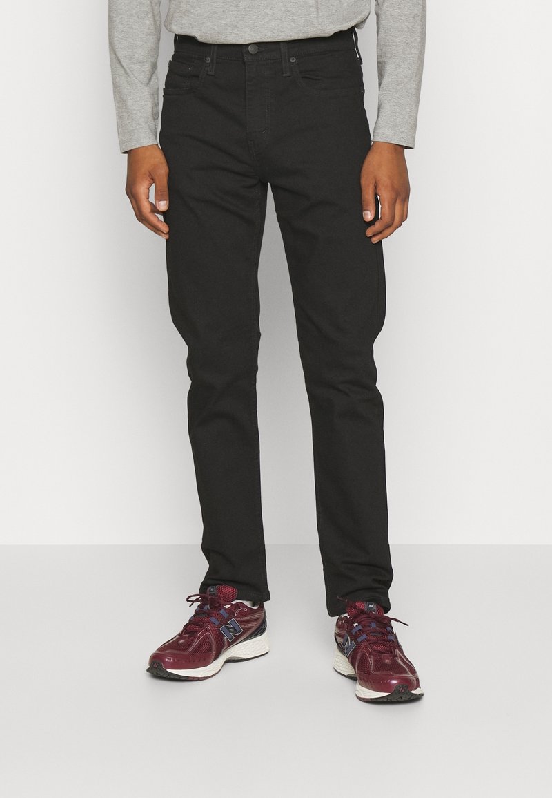 Homme portant un jean slim noir, une chemise grise à manches longues et des baskets New Balance rouge foncé, debout devant un fond blanc.