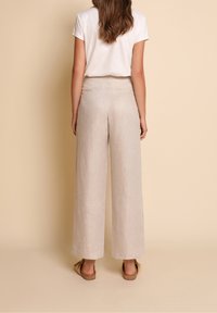 Kvinna som står med ryggen mot kameran iförd beige vida byxor, vit kortärmad topp och bruna platta sandaler mot en beige bakgrund.