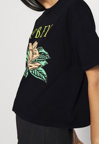 Černé bavlněné tričko s květinovým grafickým designem v zelené a béžové barvě a textem. Krátké rukávy a volný střih, s rovním lemem.