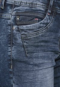 Jeans en denim dans une teinte bleu foncé délavé, avec une poche avant, des coutures décoratives et un bouton en métal près de la taille.