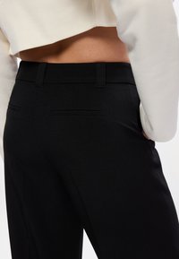 Melnas piegriezuma bikses ar gludu tekstūru, kas aprīkotas ar aizmugurējām kabatām un pieguļošu jostasvietu. Augšējā daļa ir daļēji pārklāta ar vieglu džemperi.