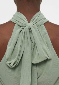 Σκοτεινόχρωμο άτομο που φοράει ανοιχτό πράσινο τοπ με λαιμόκοψη halter δεμένο με μεγάλο φιόγκο πίσω από τον αυχένα, αποκαλύπτοντας το πάνω μέρος της πλάτης και τον αυχένα.