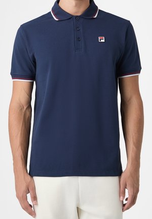 Homme portant un polo bleu marine avec un bord rayé rouge, blanc et bleu marine sur le col et les manches, ainsi qu'un petit logo sur la poitrine gauche.