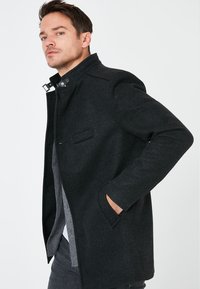 Abrigo de mezcla de lana negro con cuello alto, bolsillos laterales y sutiles detalles de costura. Corte entallado con una textura suave.