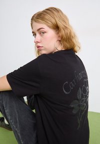 Crna pamučna majica kratkih rukava s cvjetnim uzorkom i imenom branda "Carhartt" na leđima. Model sjedi, nosi sive traperice.