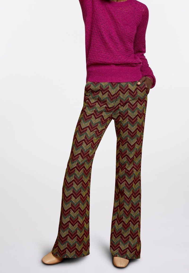Femme portant un pull en maille bordeaux et un pantalon à jambes larges à motifs en zigzag aux tons rouges, verts et jaunes, debout avec une main dans la poche.